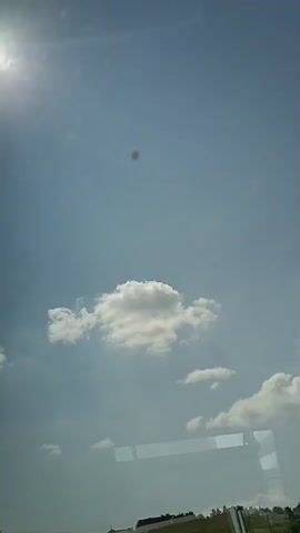 The video shows a view from inside a vehicle looking out a window. The sky is blue with scattered white clouds. There is a dark spot in the sky that appears to be a flaw on the window or a very distant object. Below the sky, there are green hills with some houses and trees. There is also a black car parked in a driveway next to one of the houses. Laundry is hanging on a clothesline in the yard of one of the houses. Reflections of the interior of the vehicle are visible on the window. The video is shot from a moving vehicle, as indicated by the subtle shift in perspective and the slight blur of distant objects.