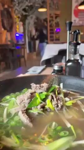 The video shows a close-up of a steaming bowl of soup, likely ramen, topped with sliced beef and scallions. The soup sits on a wooden table in what appears to be a restaurant with Japanese-style decor. In the background, a person is walking, tables and chairs are visible, and there's a decorative tree branch. A bottle with a pump dispenser and a small jar stand on the table next to the bowl.