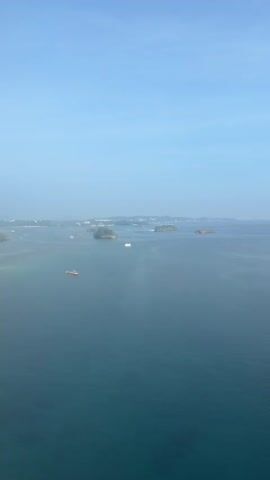 This aerial drone footage captures a serene seascape under a clear blue sky. The camera pans slowly over a vast expanse of tranquil, turquoise water dotted with several small, verdant islands. A few boats, including a larger cargo vessel and smaller white motorboats, are visible navigating the water, creating gentle wakes. The water near the islands appears shallow and clear, revealing a rocky or coral seabed. In the distance, a hazy coastline with some structures and lush greenery can be seen. The overall impression is one of peaceful isolation and natural beauty.