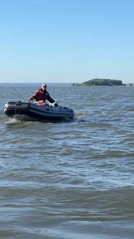 A person is in a small inflatable boat on a body of water. The person is wearing a red jacket and a hat and appears to be fishing with a fishing rod. The boat has an outboard motor. The water is choppy and there is a shoreline with trees in the distance. The sky is clear and blue.