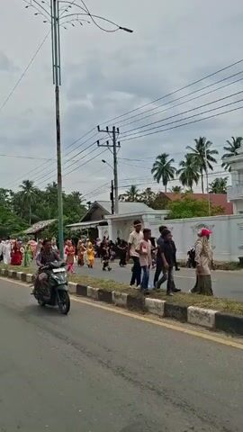 The video shows a street scene with several people walking and riding motorcycles. There is a procession of people in colorful traditional clothing walking along the side of the road. Some are carrying umbrellas or banners. Several motorcycles are also visible, some with multiple passengers. The environment appears to be a rural or suburban area with trees and houses visible in the background. The sky is cloudy. The overall atmosphere suggests a cultural event or celebration.