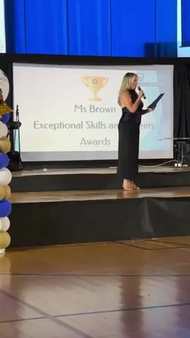 The media file shows a woman standing on a stage, holding a microphone and a piece of paper. She is wearing a black dress and appears to be speaking. Behind her, a large screen displays text that reads "Ms Brown Exceptional Skills and Talents Awards". A golden trophy icon is also visible on the screen. The stage is set up with black curtains and some equipment, possibly for a presentation or performance. The floor appears to be a polished wooden surface, typical of a gymnasium or auditorium. To the left of the stage, there is a cluster of balloons in blue, white, and gold. The overall scene suggests an awards ceremony or a similar event. The lighting indicates it is likely indoors and during an event, possibly evening. The location context is San Pedro De Alcántara, Spain.