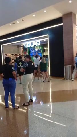 A group of people are gathered in front of a store with a sign that says "GUIAQ". The store appears to be a retail establishment. A man in a white shirt and camouflage shorts is clapping his hands in the air, facing a screen displaying images. Several other individuals are watching him, some with their hands raised. The environment appears to be a shopping mall with polished floors and a bright ceiling.