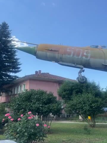 A military jet fighter airplane, painted yellow, red, and green, with the number 509 visible, is on display outdoors. It is mounted on concrete supports that elevate it above ground level. The jet is angled upwards, with its nose pointing towards the left of the frame. In the background, there is a pink building with a red-tiled roof, several green bushes and trees, and a clear blue sky. Some rose bushes with pink flowers are in the foreground to the left, and a white car is visible on the right side of the frame behind some bushes. The ground is covered in grass with some white flowers.