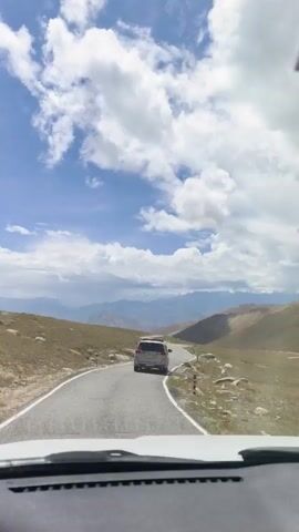 The video shows a point-of-view shot from inside a vehicle traveling on a paved mountain road. In front, a silver SUV is driving ahead on the winding road. The landscape consists of barren, brown hills on either side of the road, with distant, hazy mountains visible under a partly cloudy blue sky. The weather appears to be sunny.