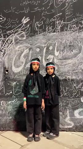 Two young girls stand side by side facing the camera in front of a dark wall covered in white writing. Both girls are wearing black headscarves and black shirts. The girl on the left has "online kids" written on her shirt in green and silver. Both girls are wearing headbands with the colors of the Iranian flag: green, white, and red. The girl on the left is wearing a black scarf tied around her neck, and the girl on the right is also wearing a black scarf tied around her neck. They appear to be standing still. The writing on the wall behind them is in a script that appears to be Arabic or Farsi. The overall lighting is dim, with some illumination coming from the front, casting shadows on the wall.