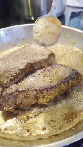 The video shows two strips of seasoned steak cooking in a frying pan. A spoon is used to baste the steaks with the pan juices. The steaks are seared on both sides and appear to be nearing completion. The pan is made of stainless steel and is filled with bubbling butter and pan drippings. A pot is partially visible in the background. The lighting is dim and appears to be artificial.