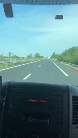The video shows the interior of a vehicle, looking out through the windshield at a highway. The dashboard is visible, with a phone mounted on the left side displaying a navigation app. The car's radio is on, with an orange display. The highway stretches into the distance, with white lines marking the lanes. On either side of the highway, there are guardrails and green vegetation. The sky is clear and blue. The video appears to be a first-person view from the driver's seat, suggesting the vehicle is in motion.