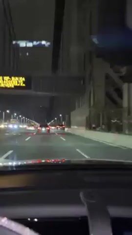 A video, shot from the dashboard of a vehicle, records the view of the George Washington Bridge at night. The bridge's support cables are visible above the roadway. A few cars are visible on the bridge, driving away from the viewer. The windshield is tinted, and the interior of the vehicle is dimly lit.
