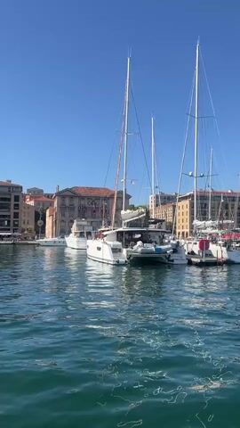 This video displays a harbor on a clear day. Several sailboats, including a catamaran, are docked in the foreground. In the background, a building with a red tiled roof and flags is visible, surrounded by other buildings. The water is a greenish-blue hue, reflecting the sky and the boats. No people are clearly discernible in the video.