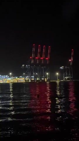 A nighttime shot of a large industrial port, likely a container terminal, is seen from the water. Multiple towering cranes with red lights at their tops dominate the background, illuminated against the dark sky. Below the cranes, rows of stacked shipping containers are visible, also lit by industrial lighting. The lights from the port reflect and shimmer on the dark, rippling surface of the water in the foreground. The scene conveys a sense of industrial activity continuing into the night.