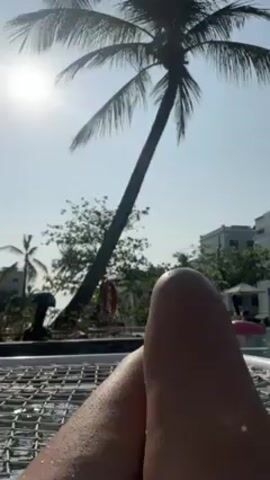 The video shows a low-angle view of a person reclining in what appears to be a hammock-style lounge chair, with their legs visible in the foreground. Behind them is a scene with palm trees silhouetted against a bright, sunny sky. In the middle ground, there are other trees, buildings, and potentially pool-related objects like a life preserver. In the background, it's possible to discern what appears to be a building structure. The environment suggests a tropical or resort-like setting.