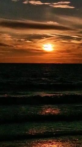 A time-lapse video shows a sunset over the ocean. The sun, a bright orange orb, is partially obscured by dramatic orange and gray clouds. The ocean below is dark, with gentle waves creating ripples and reflections of the sunset. The colors transition subtly from brighter oranges near the horizon to darker hues as the eye moves upwards, indicating the passage of time as the sun sets.