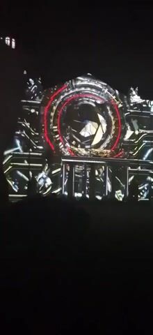 The video displays a nighttime projection mapping show on a building. A large, circular, glowing blue orb is centered on the facade, surrounded by a red ring. Intricate, mechanical-looking designs in yellow and white light are projected across the rest of the building's surface, resembling circuitry and pipes. Silhouettes of people are visible in the foreground, looking up at the display. The overall effect is futuristic and elaborate.