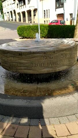 This is a close-up, vertical video of a stone fountain with water flowing out of a metal pipe. The stone fountain is round and textured to resemble wood. Inscribed on the fountain are the numbers "806 - 2006" and "1200 JAHRE FUERZ". The fountain is situated in an outdoor environment with a paved walkway made of bricks visible in the foreground and right side of the frame. Behind the fountain, a manicured green hedge lines a street, and buildings with windows and balconies are visible. A red car is parked near one of the buildings. The lighting suggests it is daytime, possibly late afternoon, with shadows cast by the fountain and walkway. The water from the fountain splashes into a basin filled with water.