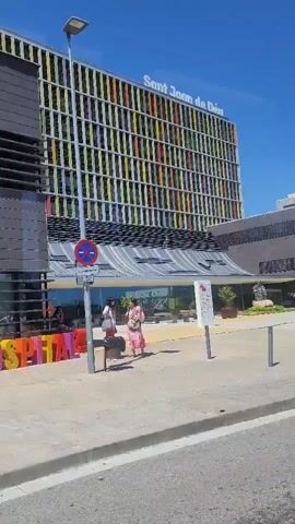 The video shows the Sant Joan de Déu hospital in Barcelona, Spain. The building is a large, modern structure with a colorful facade. There are a few people walking in front of the building, and a scooter is parked nearby. The sky is clear and blue.