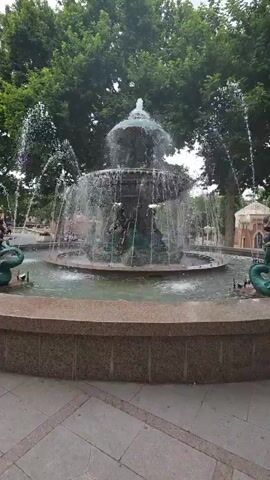 The video shows a close-up of a tiered fountain with water spraying from multiple spouts. The fountain is decorated with what appear to be mythical sea creatures, possibly mermaids or sea serpents, and has a multi-tiered design. The water falls into a circular basin at the bottom. The fountain is surrounded by a stone-like base, and beyond that, a paved area. In the background, there are lush green trees with a canopy overhead, and some structures that appear to be buildings or gazebos. The lighting suggests it is daytime. The camera pans slightly from left to right, providing a 360-degree view of the fountain and its immediate surroundings. There are no visible people actively interacting with the fountain in the foreground.