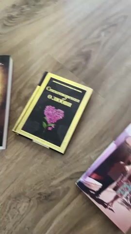 The video shows a collection of books laid out on a wooden floor. The books appear to be of various sizes and genres, with some having colorful covers and others more subdued designs. One book prominently displays the word "MAGIA" in large, bold letters. Another has a floral design with the word "Счастье" visible. There's also a book with "The Secret" logo and images of a sunset and a boat. Another book has neon pink and green lettering and appears to be titled "МУЖЧИНА". The camera pans across the books, sometimes with a slight blur, suggesting it might be handheld or the camera is moving. No people are visible in the frame, and the focus is solely on the books and the floor.