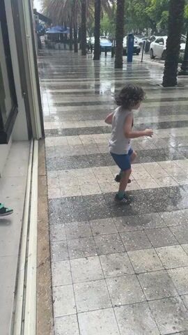 The media file shows two young children playing on a wet, tiled sidewalk during a rain shower. The child on the left, wearing a dark blue t-shirt and black shorts, is in mid-air, jumping with their legs extended. They are wearing green and black sandals. The child on the right, wearing a light grey t-shirt and blue shorts, is facing away from the camera and appears to be either jumping or about to jump as well. They are wearing dark sandals.

The sidewalk is wet and reflects the surroundings, indicating it has been raining. The ground is tiled with a pattern of light and dark gray squares. In the background, palm trees line the sidewalk, and there are buildings and parked cars visible. The scene appears to be outdoors, likely on a city street. The weather is clearly rainy, with visible water on the ground and potentially falling from the sky. The children appear to be enjoying themselves, despite the rain, as indicated by their energetic movements. The overall mood seems playful and carefree. There is no visible text in the media file.