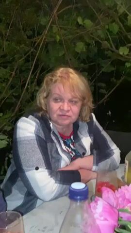A medium close-up video shows a middle-aged woman with blonde hair sitting at an outdoor table. She is wearing a checkered shirt over a patterned shirt. Her arms are crossed, and she is looking directly at the camera with a neutral expression. In the foreground, a plastic bottle and two glasses with red liquid and fruit are visible. In the background, greenery and branches are present. The lighting suggests it is evening or nighttime.