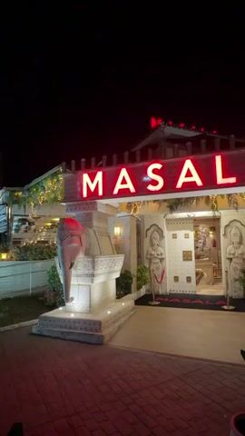 The video shows the exterior of a restaurant named MASALA at night. The restaurant has a red neon sign that spells out the name. There are two elephant statues flanking the entrance, one on each side. The entrance itself is decorated with statues of people and intricate carvings. There are also string lights and potted plants around the entrance. The ground in front of the restaurant is paved with red bricks. The overall impression is that of an Indian-themed restaurant.