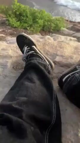 The video begins with a shot of a person sitting on a rocky cliff overlooking the ocean. The person is wearing black Vans shoes and dark jeans. The waves are crashing on the shore. The camera pans up to show the sky, which is a mix of blue and orange. The camera then moves to a person with long blond hair and wearing a purple shirt. The person smiles and makes the shaka sign with their hand.