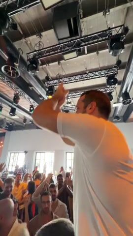A person wearing a white t-shirt is being carried on the shoulders of another person. The person on shoulders has their right arm raised in the air, waving to a crowd. The crowd appears to be at a party or event, with many people visible and raising their hands. There are lights and a truss system overhead, suggesting an indoor venue with entertainment. The overall mood seems celebratory and energetic.