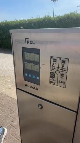 The video shows a close-up of a PCL AutoAir tire inflation machine made of stainless steel. The machine has a digital display and control buttons. A blue coiled hose is connected from the machine to the tire of a white Fiat 500 car. The car is parked on a paved surface, possibly a parking lot or gas station, with a green hedge and a utility pole visible in the background under a clear sky. The scene appears to be outdoors during the daytime. The tire inflation process is underway, as indicated by the hose connection.