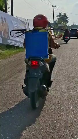 A person wearing a red helmet and a yellow shirt rides a scooter on a paved road. The person is carrying a large blue container on their back, which appears to be a sprayer. Another person on a scooter is visible in the background. There are buildings and vegetation along the side of the road. The sky is clear and the sun is shining.