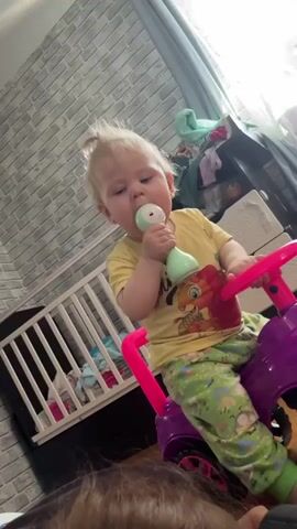 Here is a detailed description of the video:

The video shows a toddler girl sitting on a purple toy car indoors. She is wearing a yellow t-shirt with a lion design, green patterned pants, and has her blonde hair tied up in a small ponytail. The girl has a toy microphone in her mouth, then she starts holding it in her hand.

The toy car is purple with pink accents.  In the background, there is a white crib with bedding, a white dresser, and a partially visible window with a curtain. A person with brown hair is lying on the floor, partially in the foreground. The room is decorated with a brick-patterned wallpaper. Music is playing in the background.