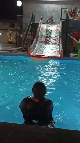 At night, a person stands in a swimming pool facing away from the camera. The person is wearing a dark t-shirt and white pants. In the background, there is a water slide with people on it. The slide is lit and the water is blue. There is a large round float in the pool. The environment appears to be an outdoor water park.