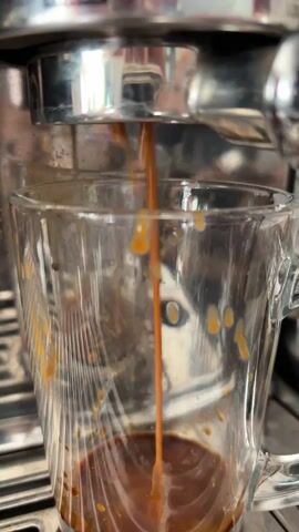 This is a close-up, vertical video of espresso being brewed into a glass mug. The espresso machine's metal portafilter is visible at the top, with a thin stream of dark brown liquid pouring from it into the mug below. The mug is clear glass, with some residue of coffee visible on the inside walls, suggesting it may have been used previously. The liquid stream is narrow and consistent, indicating a steady flow of espresso. The background is blurred but appears to be the metallic surface of the espresso machine. The lighting is moderate, highlighting the coffee stream and the glass mug. The action is solely the brewing of the espresso.