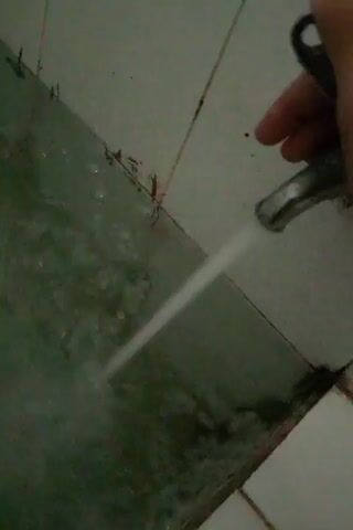 A hand is holding a faucet from which water is flowing into a green, murky pool of water. The water is creating ripples and splashes. The scene appears to be indoors, possibly a bathroom or utility area, with tiled walls visible in the background. The tiles are white and have visible grout lines and some staining or discoloration. The overall impression is one of a functional, albeit somewhat unkempt, space.
