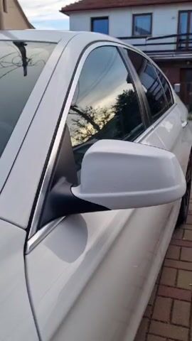 This is a close-up, side-view video of a white sedan parked on a brick driveway. The car appears to be in good condition, with a clean and shiny exterior. The reflection in the car's windows shows trees and a cloudy sky, suggesting it is daytime, possibly late afternoon or early evening given the lighting. Behind the car, a two-story house with a red-tiled roof is visible. The house has multiple windows and a balcony. The overall setting appears to be a residential area. There is no visible text in the media. The video is static, focusing on the details of the car and its surroundings.