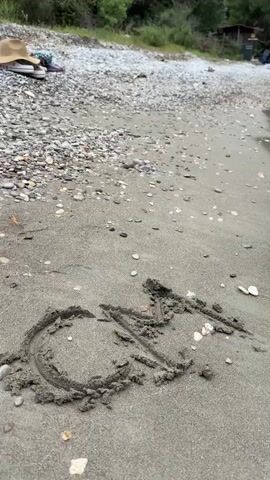 The video shows a beach scene with a sand and pebble shoreline leading to the edge of the water. In the sand, the word "SEA" is written. In the background, there is a pebble-covered section of the beach, where a hat and some clothing or bags are placed. Further back, there's a small wooden structure or cabin, surrounded by lush green trees and shrubbery. A mountain covered with trees and rock formations is visible in the background, partially obscured by low-hanging clouds. The sky is overcast and gray. There are waves softly washing onto the beach.