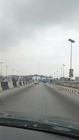 The video shows a view from inside a vehicle driving towards a toll plaza. The road is paved and has multiple lanes. There are concrete barriers on either side of the road. The toll plaza has multiple booths with signs and lights. Several cars are visible on the road, some approaching the toll booths. The sky is overcast. The video appears to have been recorded during the daytime.