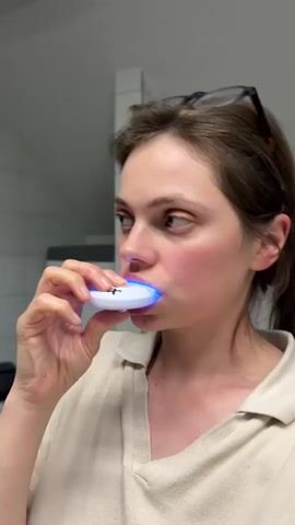 A young woman with brown hair pulled back and glasses perched on her head is holding a white dental appliance with a blue light to her mouth. The appliance is in her mouth, and the blue light is illuminated, suggesting she is undergoing a teeth whitening treatment. She is wearing a beige collared shirt. The background appears to be a bathroom, with white tiled walls and a glimpse of a white appliance in the background. The lighting is bright, typical of a bathroom.
