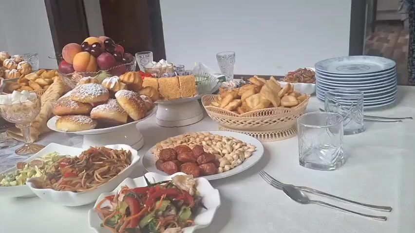 The video shows a long table laden with a variety of food and dishes. The table is covered with a white tablecloth patterned with grey swirls. On the table are several plates of food, including noodles, salads, fried dough, pastries, fruits, and what appear to be sausages and nuts. There are also stacks of plates, glasses, and cutlery. The food is arranged in an abundant display, suggesting a feast or gathering. The background is indistinct, but appears to be an indoor setting with neutral-colored walls and some furniture. No people are visible in the video. The lighting is soft and diffused.