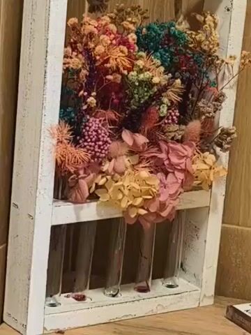 A close-up video shows a decorative flower arrangement in a weathered white wooden frame. The arrangement includes dried flowers of various colors: orange, pink, yellow, blue, green, and purple. The frame has test tubes at the bottom, some containing a small amount of red liquid. A rope handle hangs from the top of the frame. The background is a light brown wooden surface.