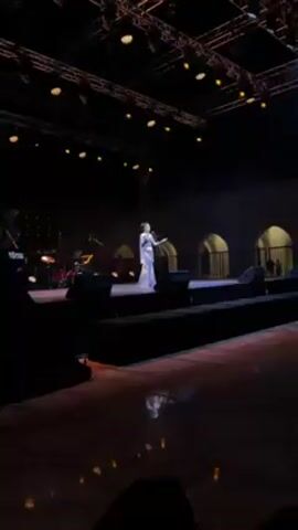 The video shows a woman singing on a stage. She is wearing a long, light blue dress with lace details and a sheer shawl. Her hair is styled up. She is standing at a microphone and gesturing with her hands as she sings. The stage is dark with spotlights on her. There is a band behind her to the left and speakers on the stage in front of her. The background features arched openings.