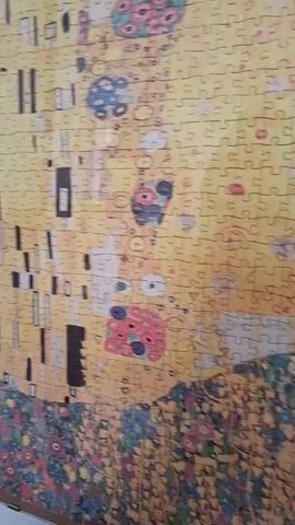 The video shows a close-up, panning shot of a completed jigsaw puzzle. The puzzle depicts Gustav Klimt's painting "The Kiss," characterized by its intricate patterns and predominantly gold and yellow hues. The image is composed of many small, interlocking pieces, and the camera moves horizontally to reveal different sections of the artwork. The foreground of the puzzle features a floral pattern with various colors including pink, purple, and green. The background is a swirling mosaic of gold and yellow, interspersed with abstract shapes and figures. The quality of the image is somewhat grainy, suggesting it was taken with a handheld device. There are no people visible in the video, nor any other objects besides the puzzle itself.