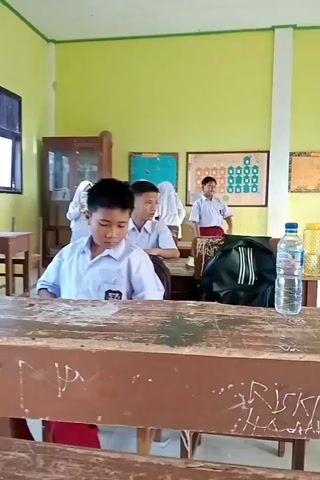 The video shows a classroom with several students wearing school uniforms. The student in the foreground is looking down at their desk. Another student in a white shirt and red pants approaches the desk with a book in their hand and places it down. A student wearing a red cap and tie stands near the back of the classroom, looking to the side. The walls of the classroom are painted green, and there are educational posters on the walls. There are desks and chairs, a backpack, and a water bottle on a desk.