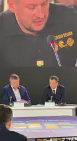 A video records an indoor event, likely a conference or presentation, from an audience perspective. The main subject is a large screen displaying a man speaking into a microphone. This man has short hair and a beard, and wears a dark polo shirt with a gold-colored insignia on his right chest area, which reads "ХЕРСОН РУСЬ" (Kherson Rus). He appears earnest as he speaks, seemingly delivering a speech in Ukrainian, with words like "гарантії" (guarantees) and "медичної" (medical) audible in the partial transcript.

Below the large screen, two men are seated at a white table, facing forward. The man on the left wears a blue suit jacket over a white shirt and has short brown hair. The man on the right is in a dark suit and tie, looking down with one hand resting near his face, possibly in thought or covering his mouth. Both men have small nameplates or documents in front of them, along with bottles or glasses on the table. In the immediate foreground, the back of an audience member's head is partially visible. The scene suggests a formal gathering in Barvinok, Ukraine, where an address is being given, possibly about the Kherson region, to an audience including the two seated individuals.