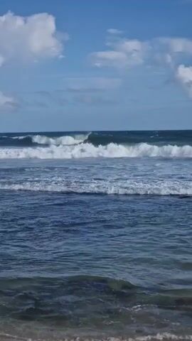 The video shows a seascape under a blue sky with scattered white clouds. The ocean is a deep blue, with waves rolling in towards the shore. The waves are capped with white foam. The beach is visible in the foreground, a sandy expanse with some rocky formations partially submerged in the water. The horizon is clear, and the overall impression is one of a sunny, breezy day at the beach.