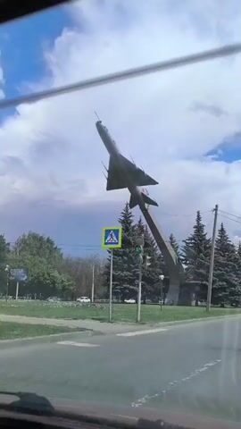 The video is shot from inside a vehicle, looking out the windshield. In the foreground, the car's dashboard and side mirror are visible. The middle ground features a street and a grassy area. Dominating the background is a monument of a fighter jet mounted on a concrete pillar, angled upwards into a cloudy blue sky. Several tall evergreen trees are positioned behind the monument. A pedestrian crossing sign and a traffic light are also visible on the left side of the road. The overall scene appears to be an outdoor public space, possibly a park or a roundabout, with elements of urban infrastructure.