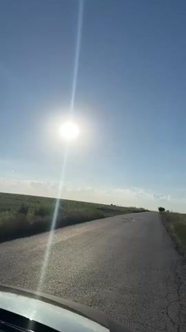 The video shows a first-person perspective of a vehicle traveling on a paved road. The road is cracked and has potholes. The sun is bright in a clear blue sky with a few scattered clouds. To the left of the road is a grassy field with some tall weeds. To the right of the road is also a grassy field. In the distance, a lone tree is visible on the right side of the road. The sun is creating a strong lens flare, obscuring some details. The video appears to be shot during the daytime.