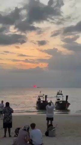 The video shows a beach at sunset. There are several people on the beach. Some of the people are standing and taking pictures with their phones of the sunset. Some are sitting down. Two boats are partially submerged in the water near the shore. The sun is visible through the clouds and is a deep red color. The sky is filled with clouds, which are shades of gray and orange. The sand is light beige. The water is calm.