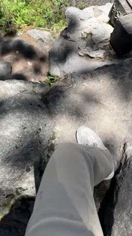 The video begins with a close-up view of a person's white sneaker and light-colored pants standing on large rocks near a flowing body of water. The camera then pans up to reveal a waterfall cascading down a rocky cliff face surrounded by lush green foliage and trees. Sunlight filters through the leaves, casting shadows on the rocks. A wooden bridge or railing is visible on the right side of the frame. The sky above is a clear, bright blue. The camera continues to pan upwards, showcasing the full height of the waterfall and the surrounding natural landscape under a sunny sky.