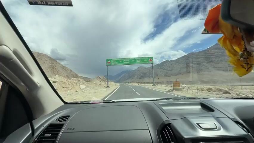 The video is a first-person view from inside a car driving on a paved road through a mountainous desert landscape. The road ahead is empty. The car dashboard and a side mirror are visible. The side mirror reflects the interior of the car and the landscape outside. A green signpost with text in English and other languages is visible ahead, indicating distances to various locations. The sky is cloudy with patches of blue. The surrounding terrain is dry and rocky, with sparse vegetation. The car appears to be moving at a moderate speed.