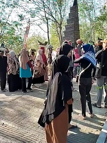 A group of people are walking in a procession outdoors. Several individuals are carrying baskets or trays of items on their heads. The people are dressed in a variety of clothing, with some wearing headscarves. There are trees in the background, and flags or banners are visible, suggesting a festive or cultural event. The ground appears to be a paved or dirt path.